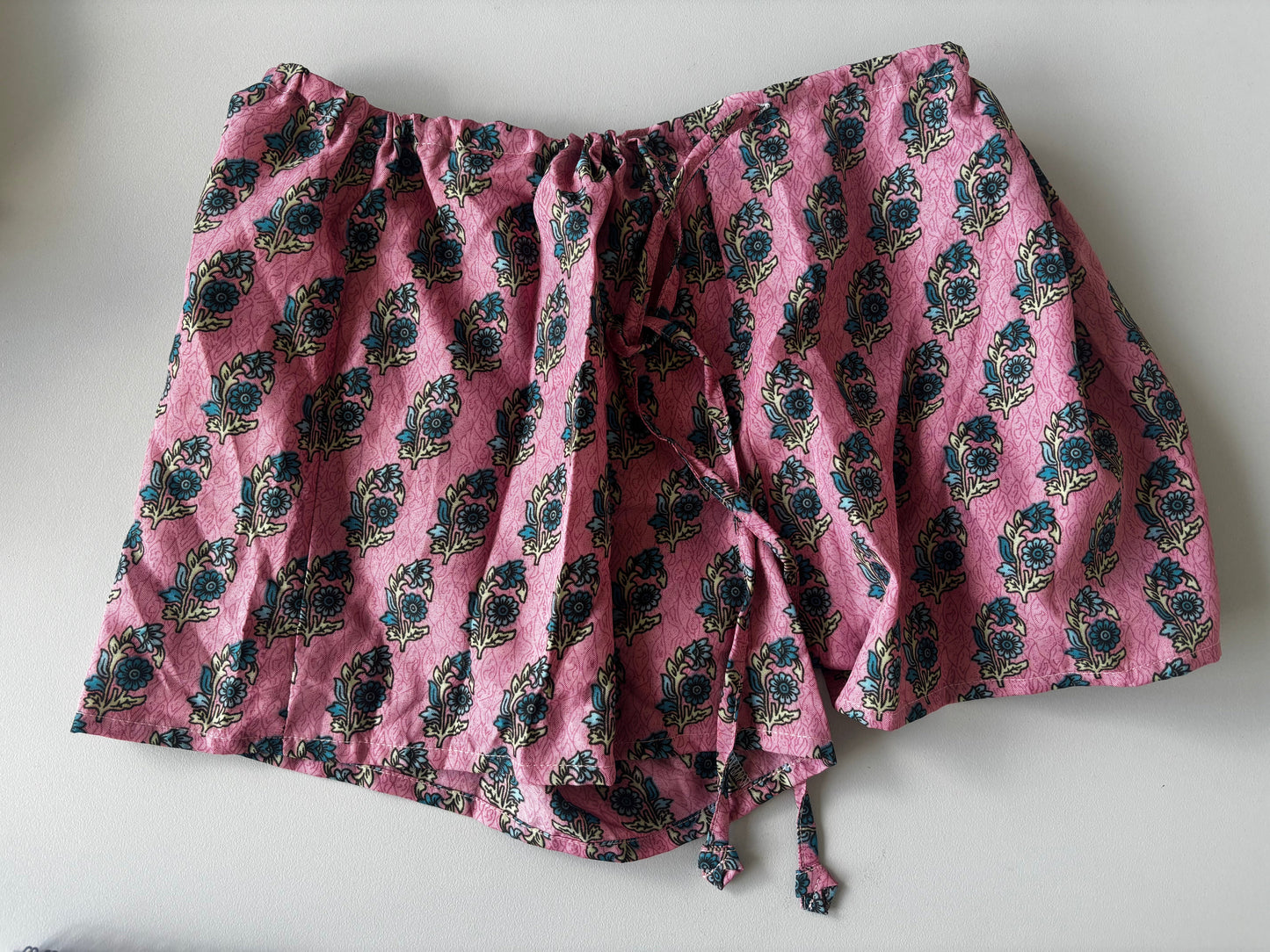A pink fabric garment, likely a skirt or dress, featuring a dark blue and green floral block print pattern and a drawstring waist, laid flat on a light gray surface.