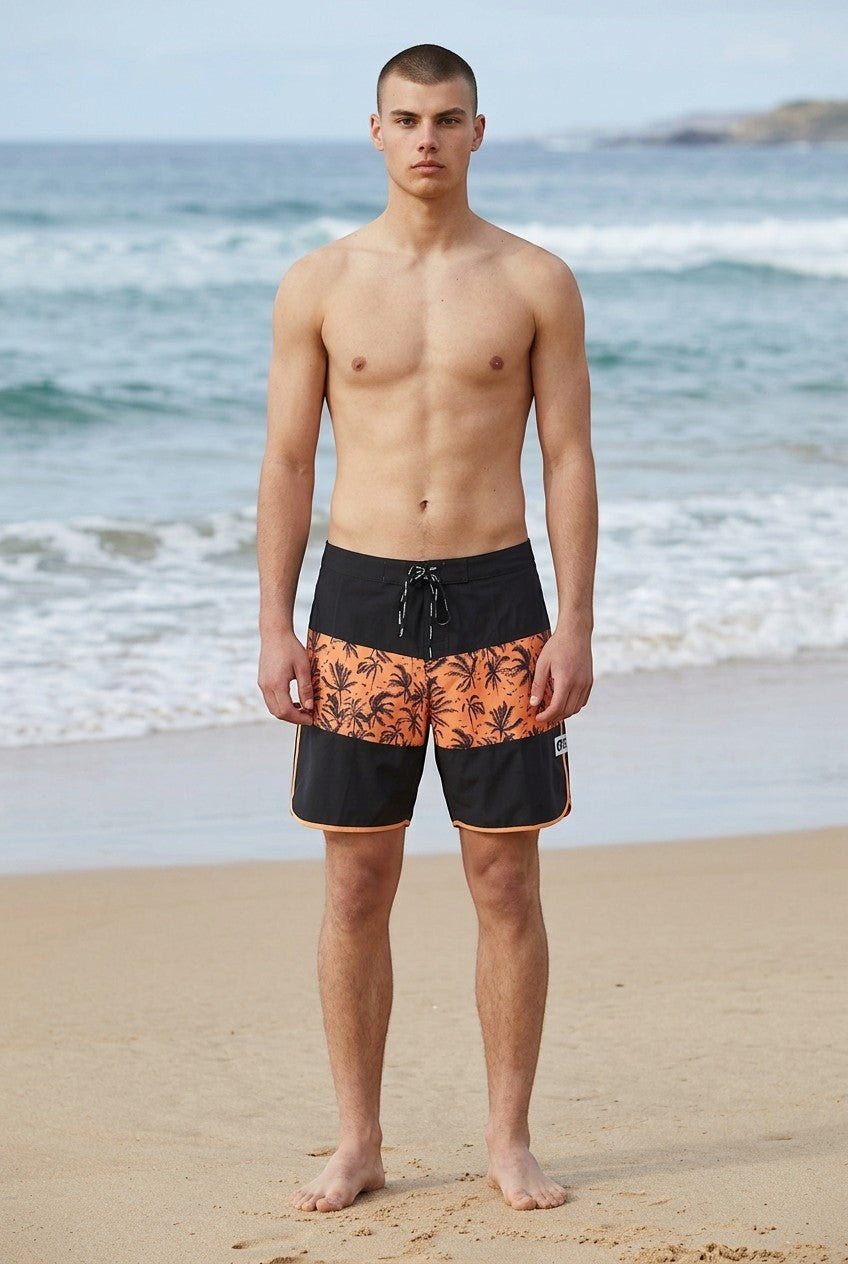 Invogue Again Andy 17 inch boardshorts in black with an orange palm tree print panel, worn by a man on the beach.