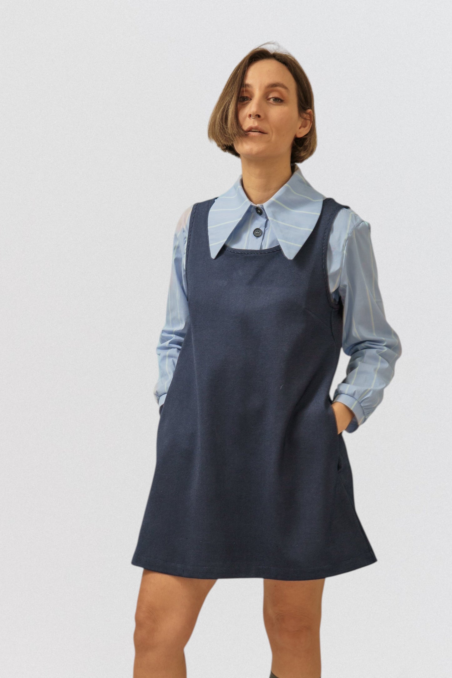 Invogue Again denim pinafore dress in dark blue worn over a light blue striped shirt with side pockets