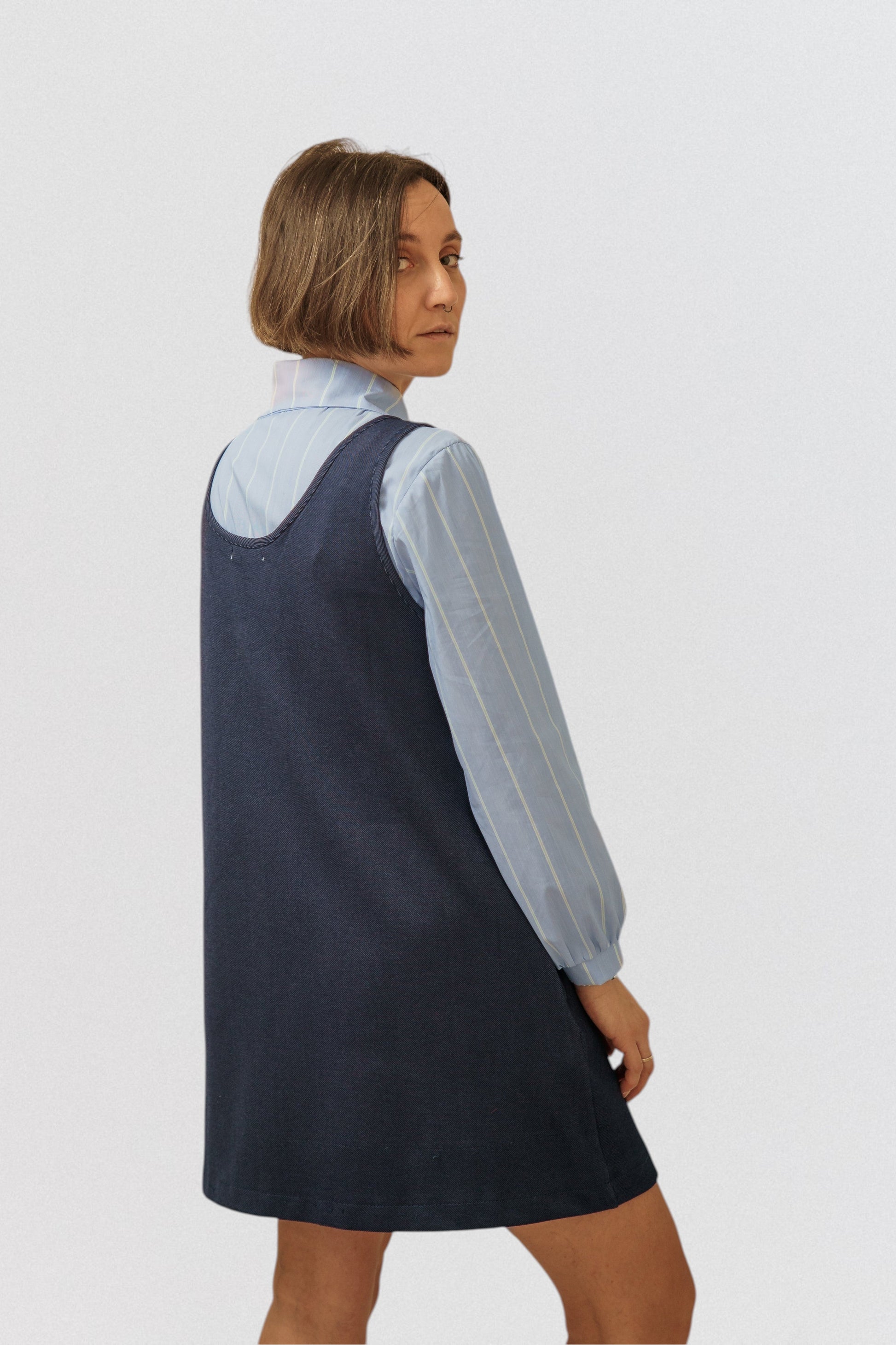 Invogue Again denim pinafore dress in dark blue worn over a light blue striped shirt by a woman, back view.
