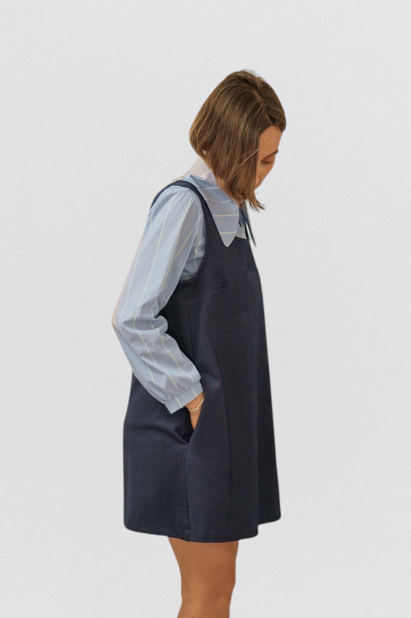 Invogue Again denim pinafore dress in dark blue with side pockets, worn over a light blue striped shirt.