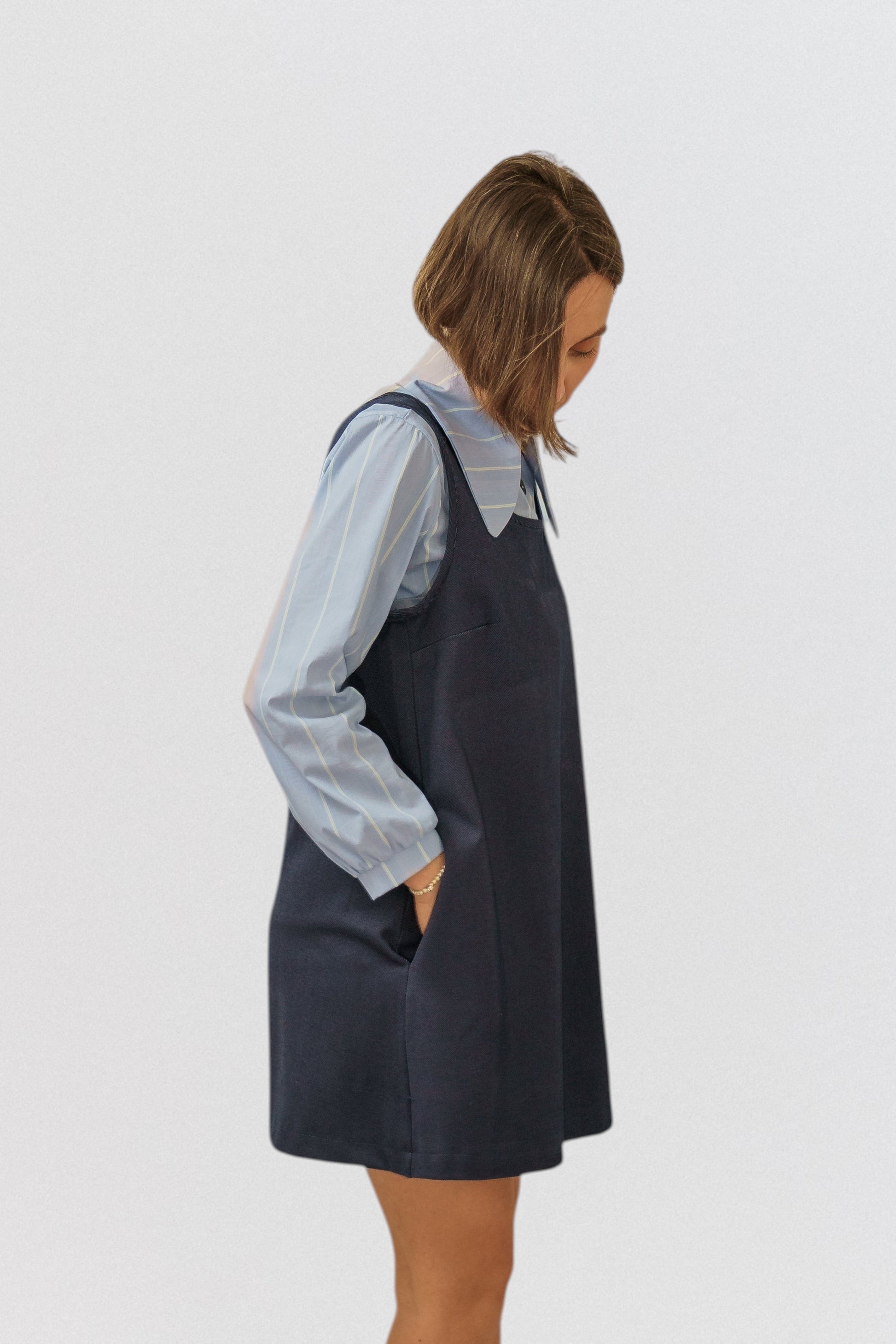 Invogue Again denim pinafore dress in dark blue with side pockets, worn over a light blue striped shirt.