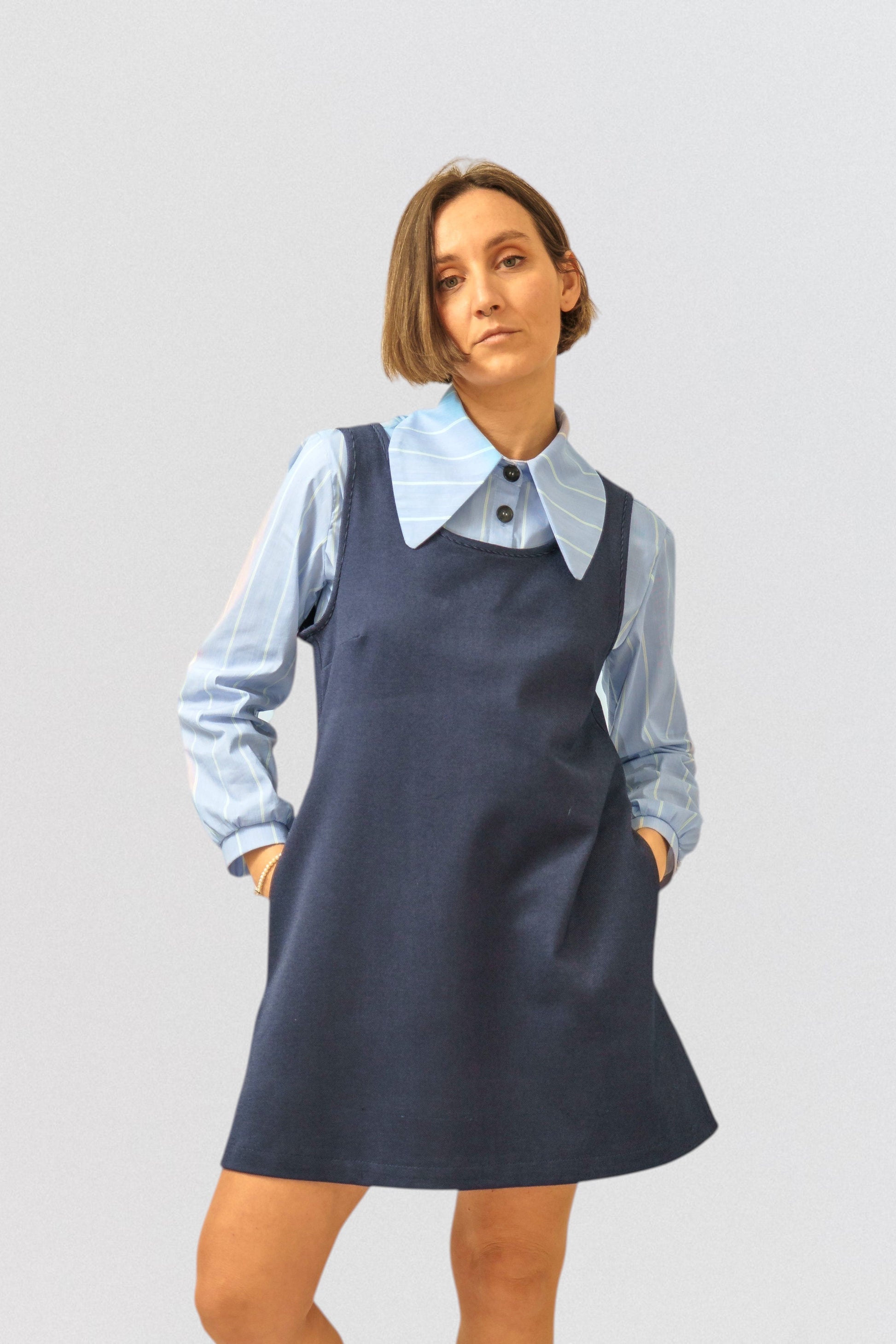 Invogue Again denim pinafore dress in navy blue worn over a light blue striped collared shirt by a female model.